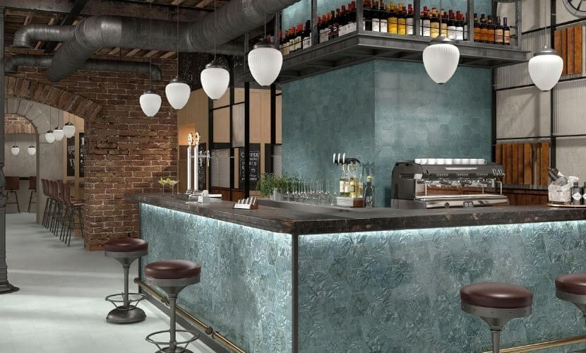 Interior de bar con mostrador de azulejos azules, taburetes y luces colgantes; ladrillos a la vista y decoración de estilo industrial.
