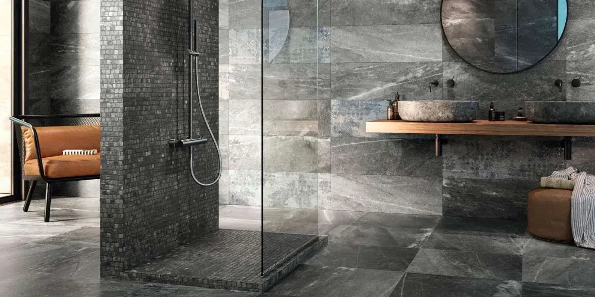 Baño con paredes y pisos de azulejos gris oscuro, ducha, espejo redondo y tocador de madera.