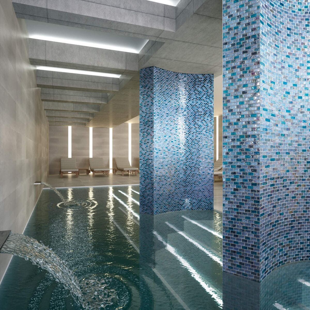 Piscina cubierta con fuente de agua, revestida con pilares y paredes de mosaico azul y tumbonas al fondo.