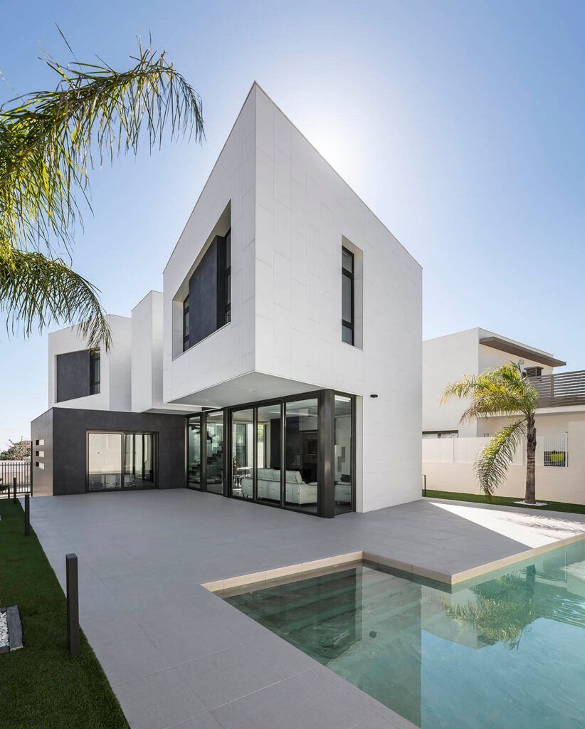 Moderna casa blanca con detalles grises, piscina y palmeras bajo un cielo soleado.