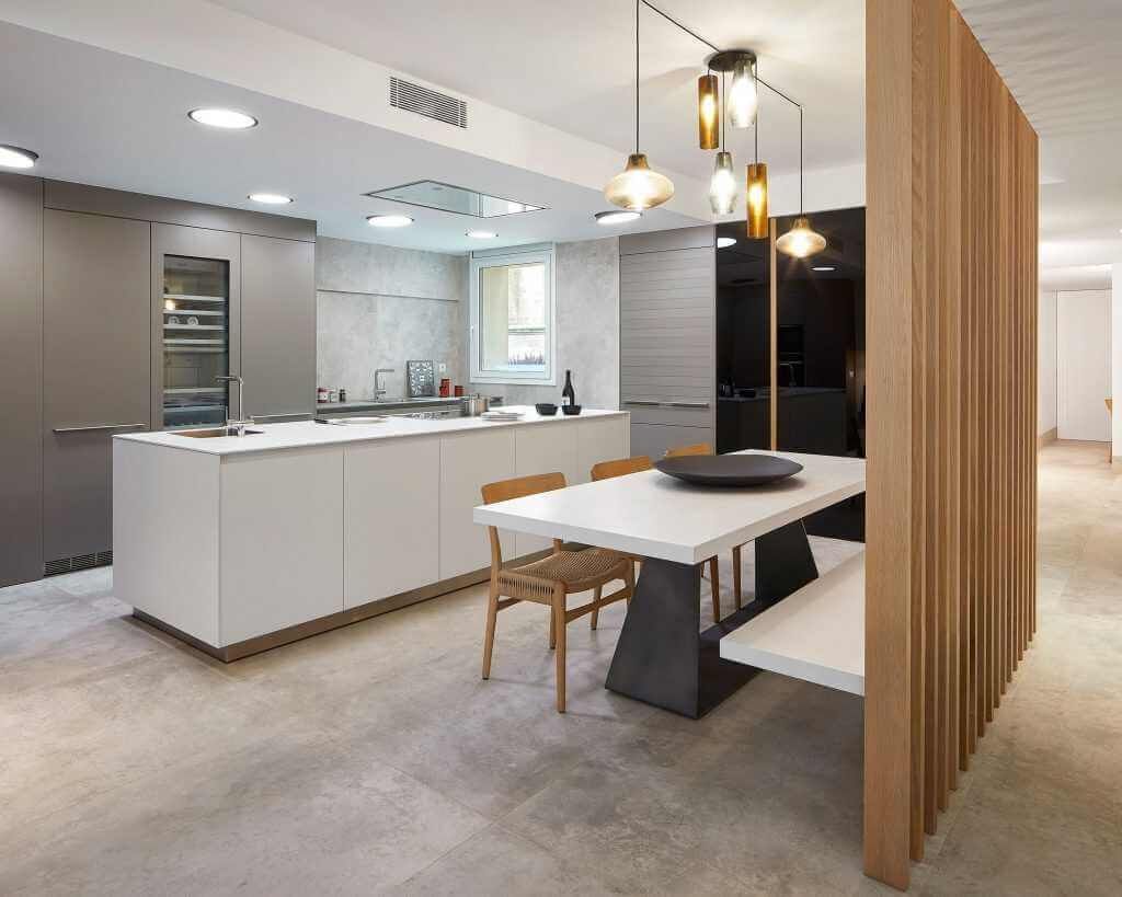 Cocina moderna con gabinetes color gris claro, isla, mesa de comedor, divisor de listones de madera y luces colgantes.