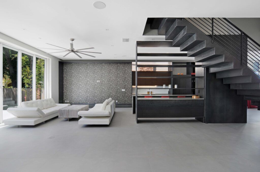 Moderna sala de estar con sofás blancos, suelo gris, escaleras gris oscuro y grandes ventanales.