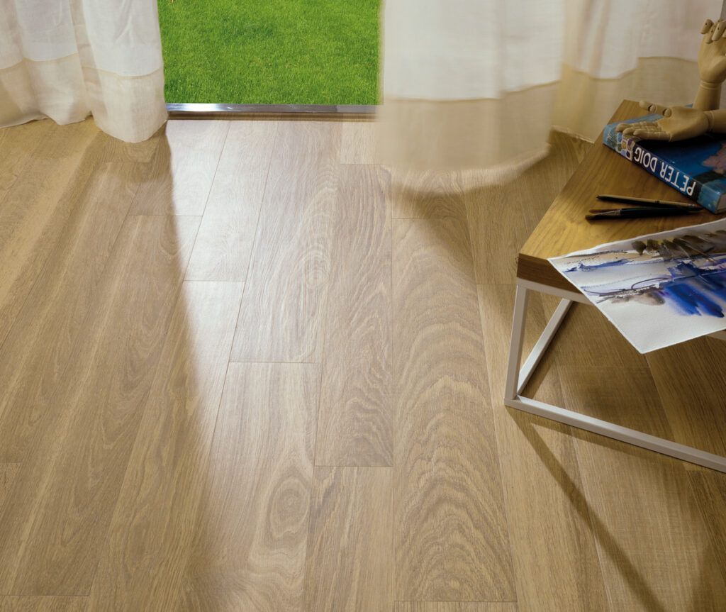 Piso de madera clara con luz solar, cortinas y una pequeña mesa con un libro y una lámina artística.