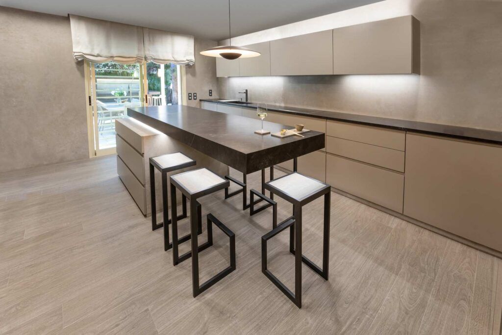 Cocina moderna con isla y taburetes, tonos neutros, elementos de madera y piedra e iluminación cenital.