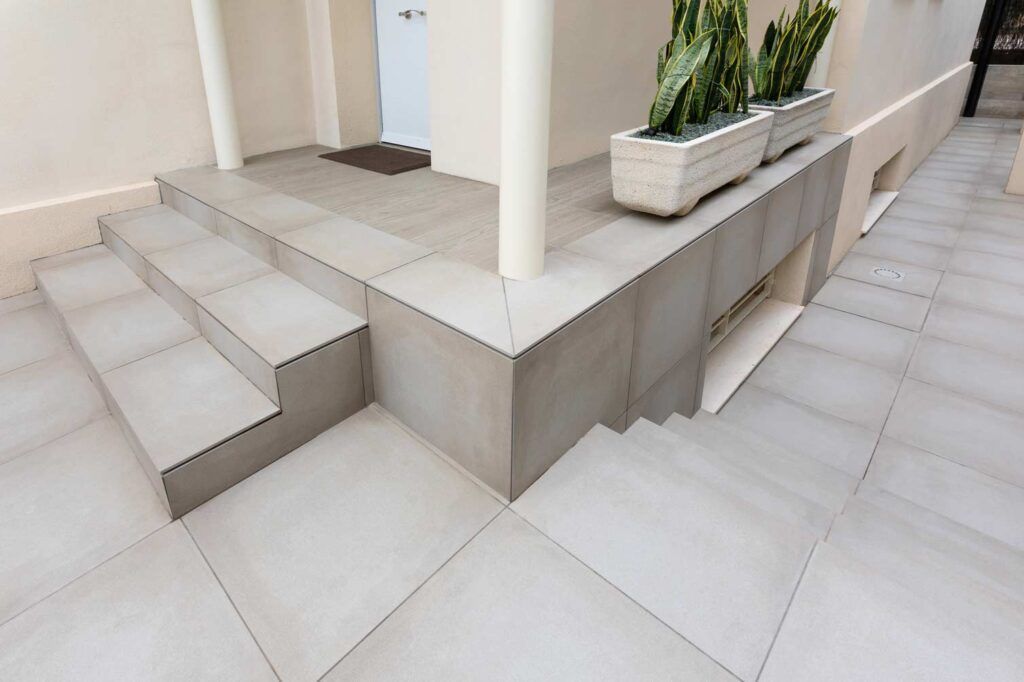 Unos escalones de hormigón conducen a un patio elevado con jardinera y columnas; tonos neutros.