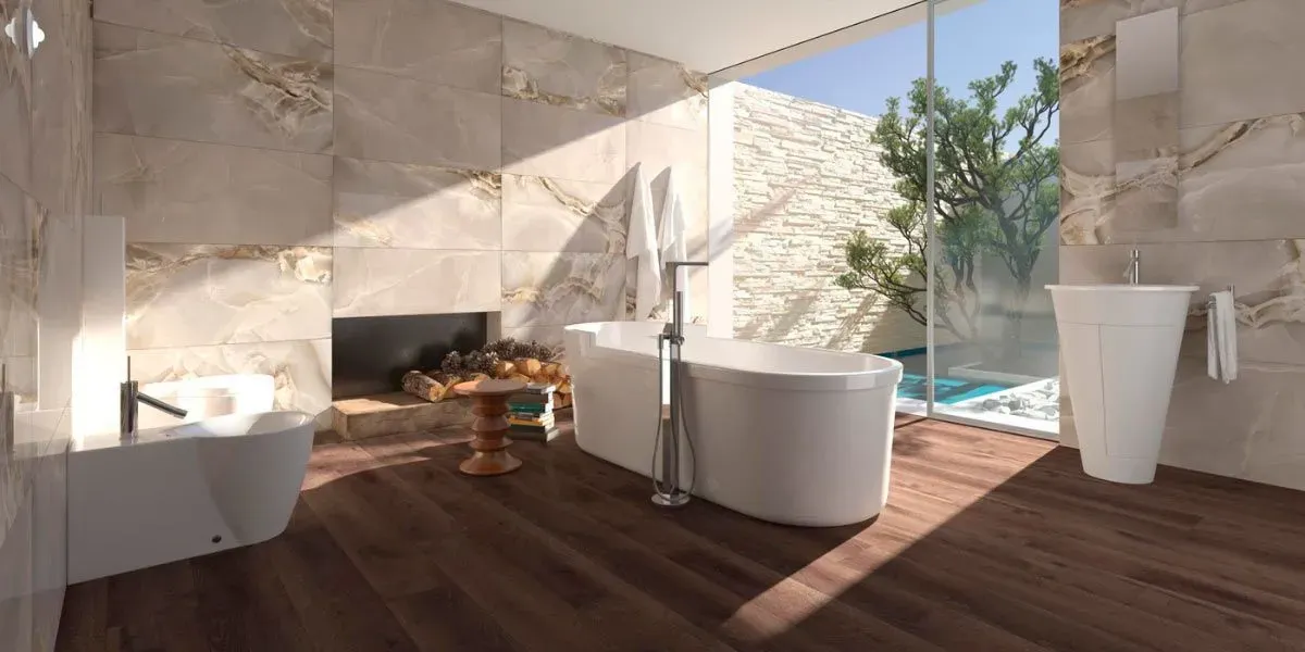 Baño con bañera, lavabo e inodoro. Un gran ventanal con vistas a la piscina y a los árboles. Suelo de madera y paredes de piedra.