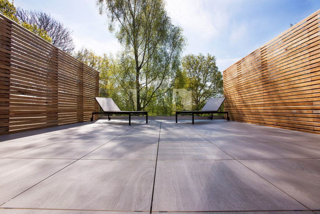 Dos tumbonas en una terraza de madera rodeada de paredes de listones de madera para mayor privacidad, con árboles al fondo bajo un cielo azul.