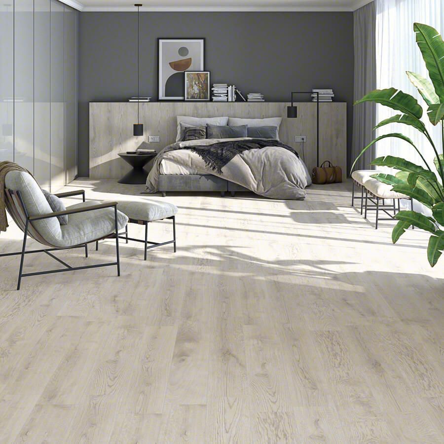Dormitorio con suelo de madera clara, cama, silla y una planta grande. Tonos grises y neutros.