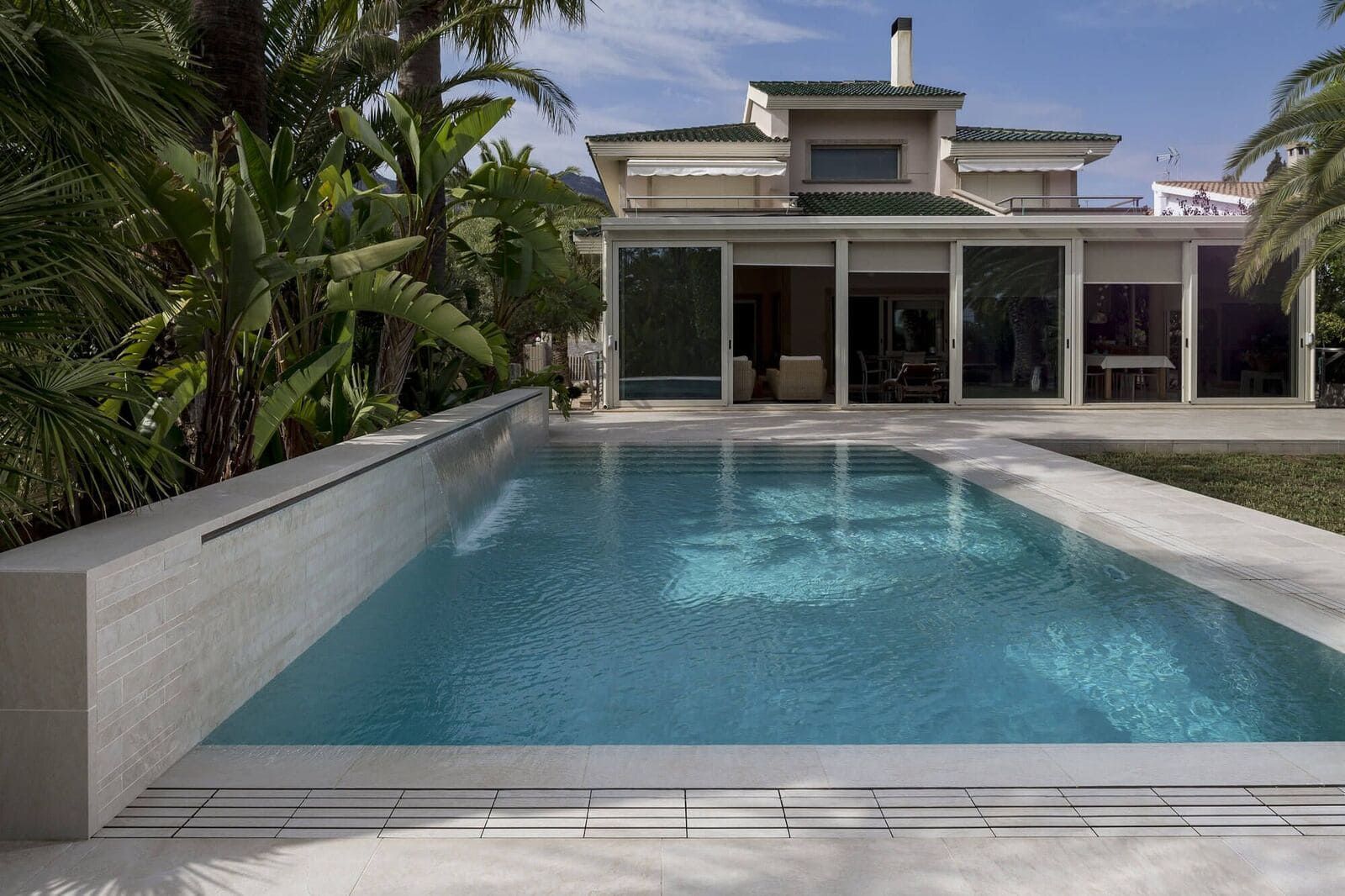 Piscina de agua azul frente a una casa moderna, con palmeras y cielo soleado.