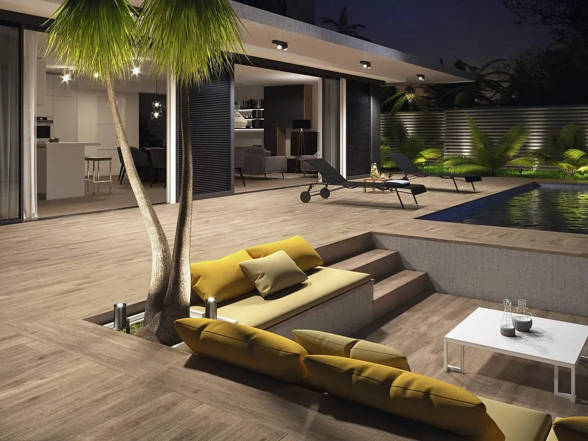 Zona de estar al aire libre con piscina, palmera y una casa moderna. Cojines amarillos y mesa de centro blanca.