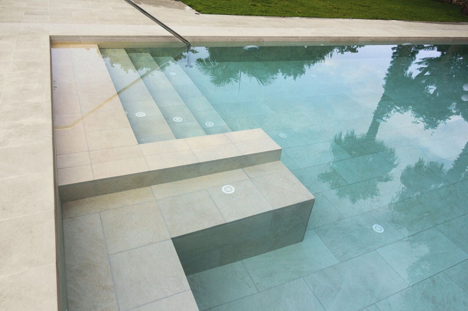 Piscina con escaleras incorporadas y azulejos de color claro. Se ven los reflejos de los árboles en el agua.