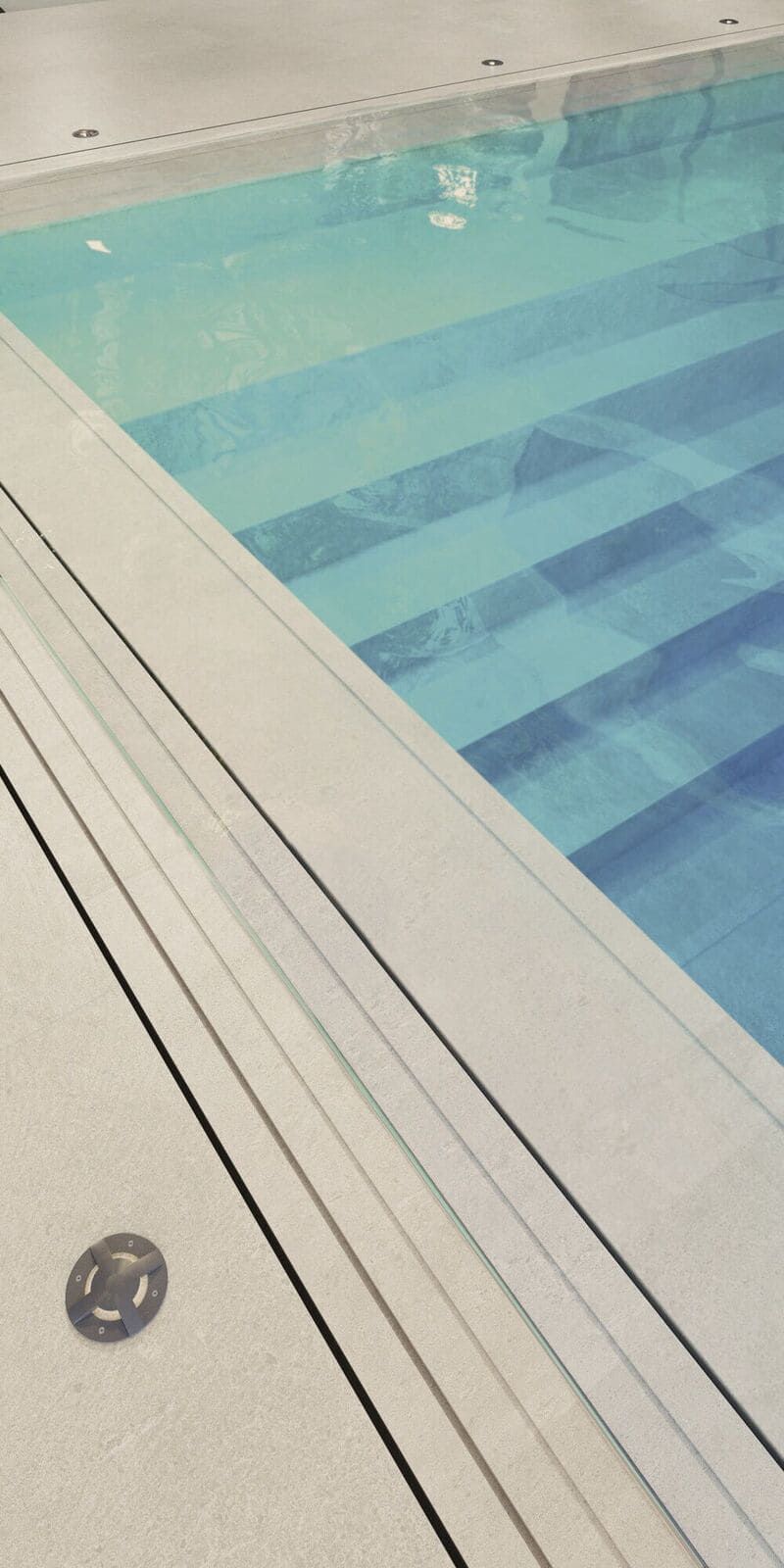 Piscina con escalones. Agua azul claro. Borde beige con líneas negras.