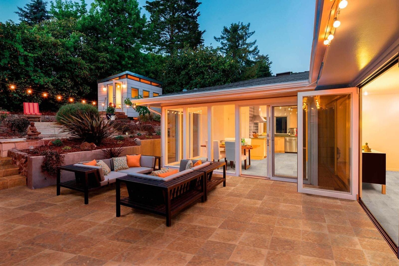 Patio con asientos al aire libre, adyacente a una casa con puertas de vidrio abiertas y luces de cadena, al anochecer.