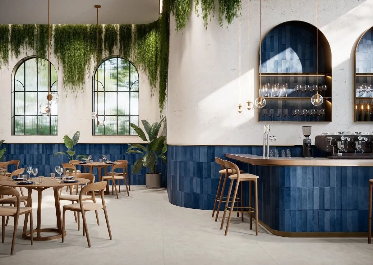 Interior de restaurante con azulejos azules, ventanas arqueadas, plantas colgantes y mesas de madera.
