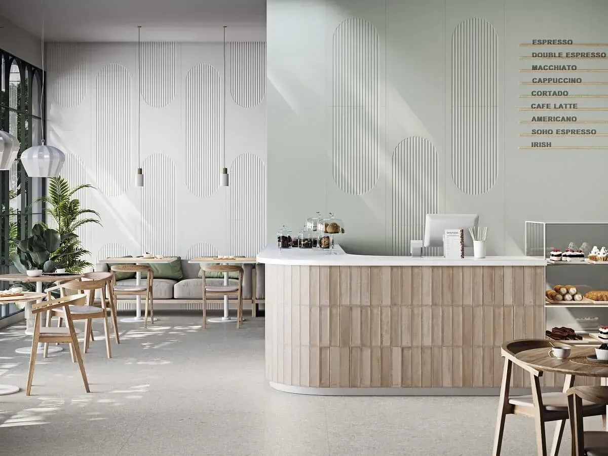 Interior de cafetería con mostrador, mesas y paneles decorativos. Tonos verde pastel y blanco, luz natural.