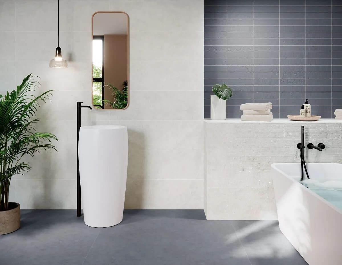 Baño con lavabo de pedestal blanco, espejo y bañera. Las paredes tienen azulejos neutros y azules, y una planta.