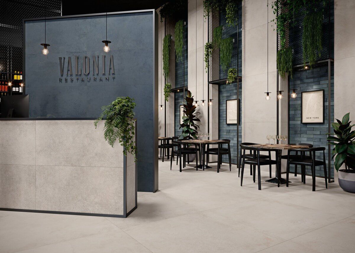 Interior del restaurante: entrada con mostrador de recepción texturizado, pared azul oscuro, mesas y sillas, plantas colgantes y luces colgantes.