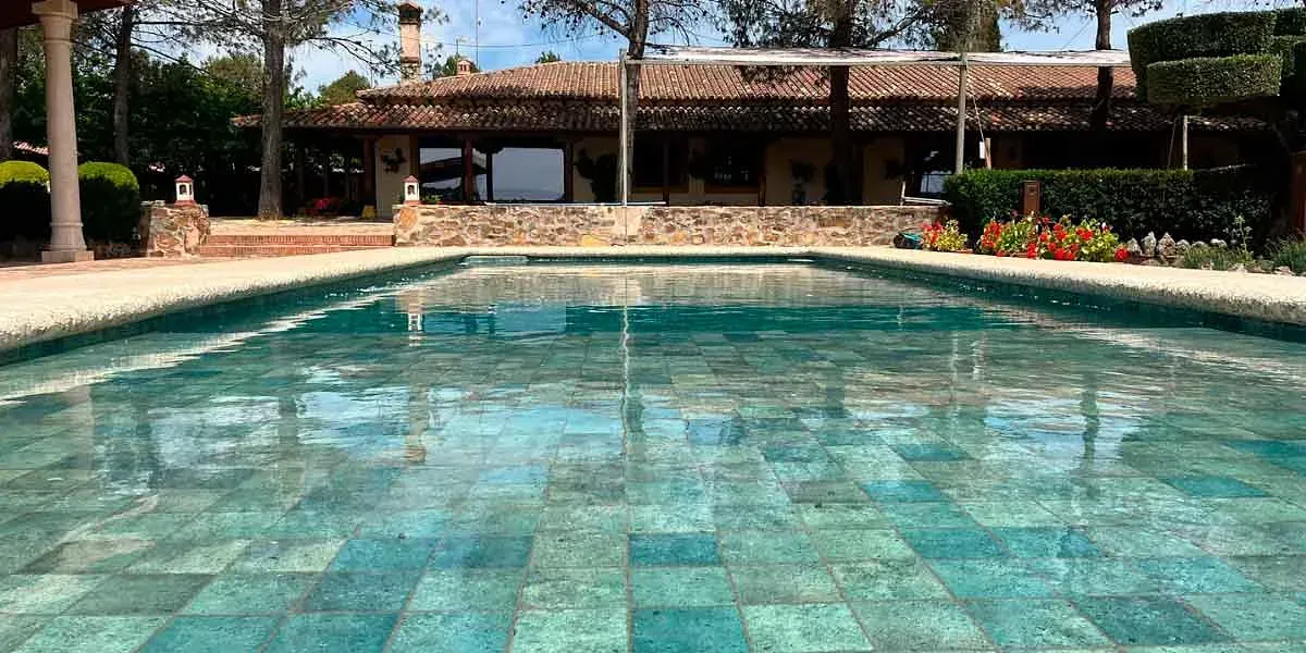 Piscina frente a un edificio con techo de tejas, árboles al fondo, agua azul verdosa.