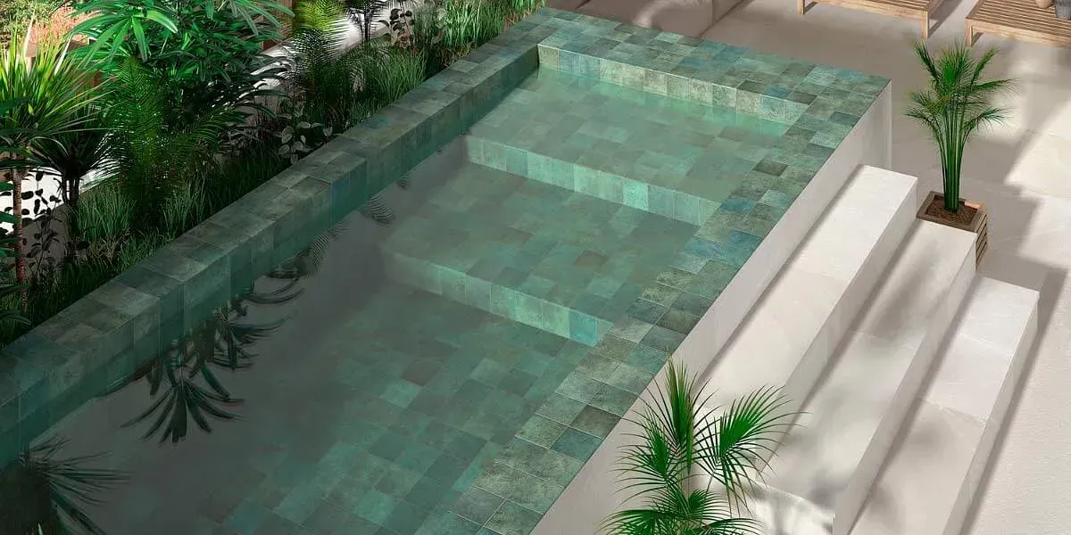 Piscina al aire libre con interior de azulejos verdes y escalones, rodeada de vegetación y escaleras blancas.