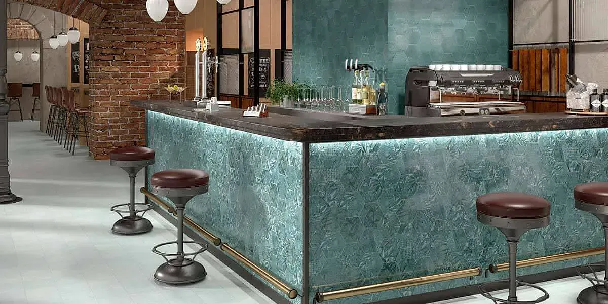 Barra con frente texturizado verde azulado, taburetes, pared de ladrillos y máquina de café expreso.