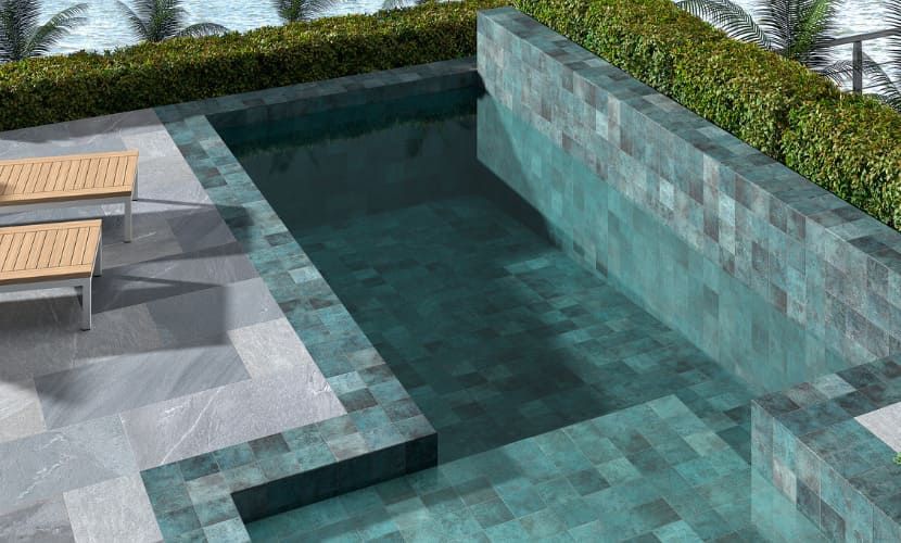 Piscina con azulejos turquesa oscuro, rodeada de un patio de piedra y un seto verde. Hay dos tumbonas a la izquierda.