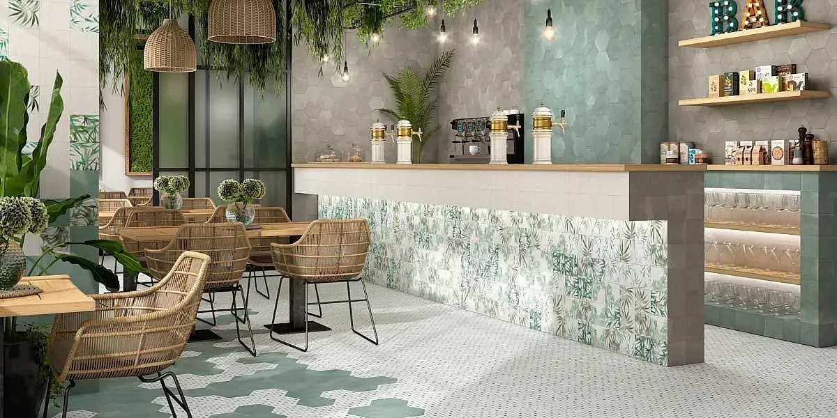 Interior de una cafetería con sillas de mimbre, plantas y una barra de color claro con azulejos estampados.