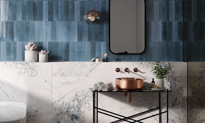 Baño con pared de azulejos azules, lavabo de cobre, espejo con marco negro y tocador símil mármol.