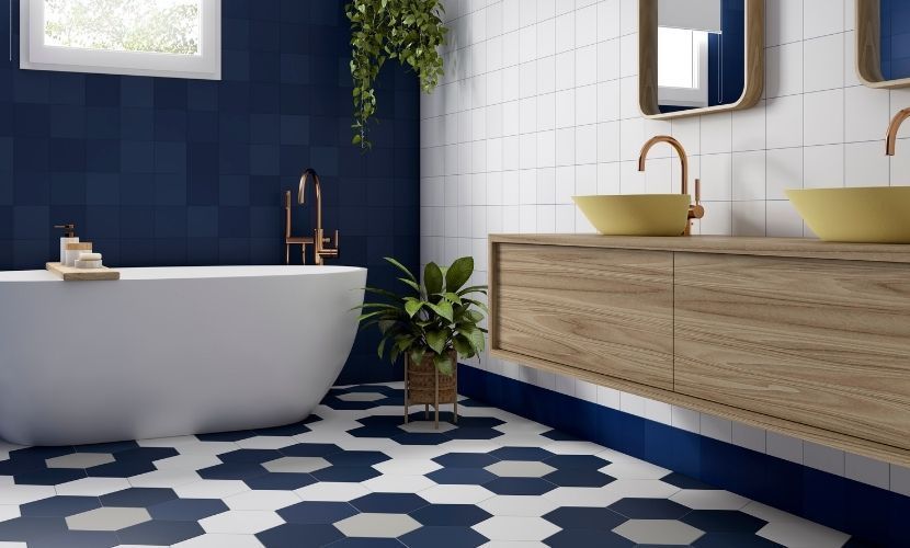 Baño con piso de azulejos florales azules y blancos, pared azul, tocador de madera y bañera blanca.