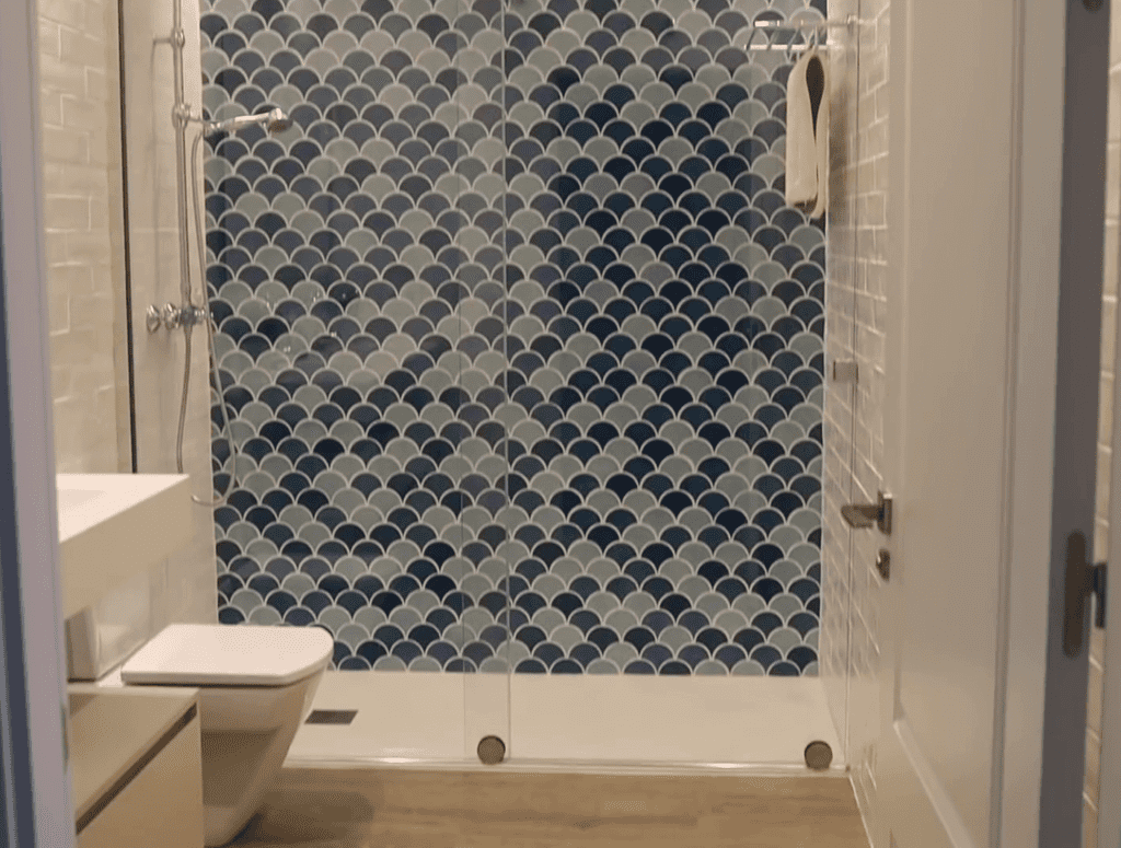 Baño con pared de ducha con estampado azul y gris, inodoro y lavabo blancos y puerta de ducha de vidrio.