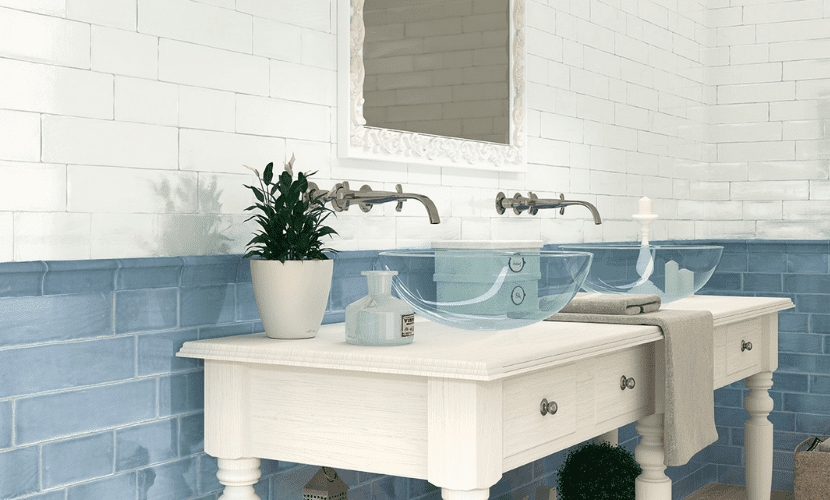 Baño con paredes de azulejos blancos y azules, tocador blanco, lavabos de cristal y una planta en maceta.