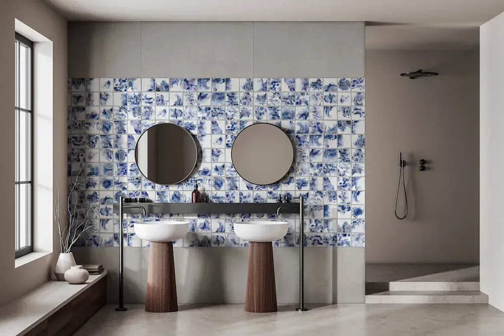 Baño con salpicadero de azulejos con dibujos azules y blancos, dos espejos redondos y lavabos con pedestal.