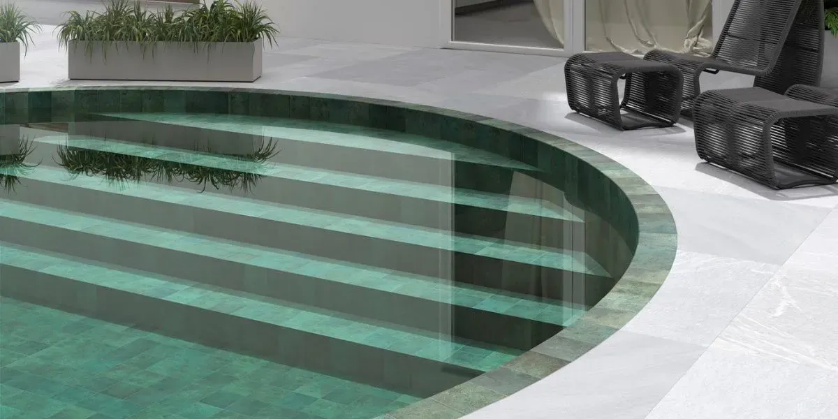 Una piscina redonda con escalones de agua turquesa, rodeada de piedra clara. Junto a la piscina hay dos sillas y una jardinera.