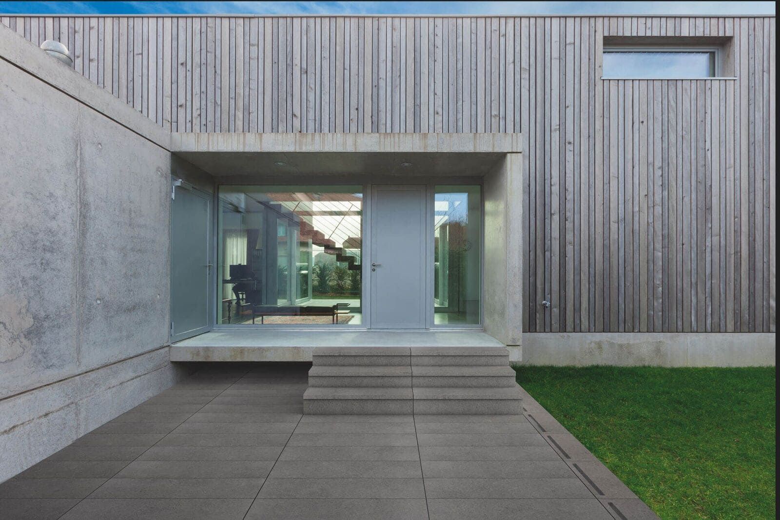 Entrada de edificio moderno con hormigón, paneles de madera y puerta de cristal. Unos pequeños escalones conducen a la puerta.