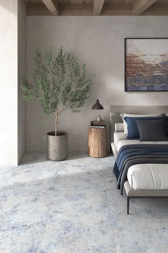 Dormitorio con suelo moteado de azul, una cama, un árbol en una maceta y un cuadro.