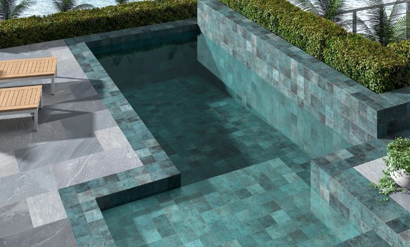Piscina de agua turquesa y paredes de baldosas, rodeada de patio de piedra gris y setos verdes.