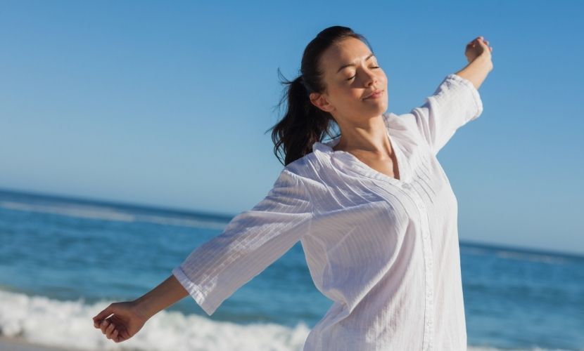 Mujer con los brazos extendidos, los ojos cerrados, sonriendo en una playa soleada.