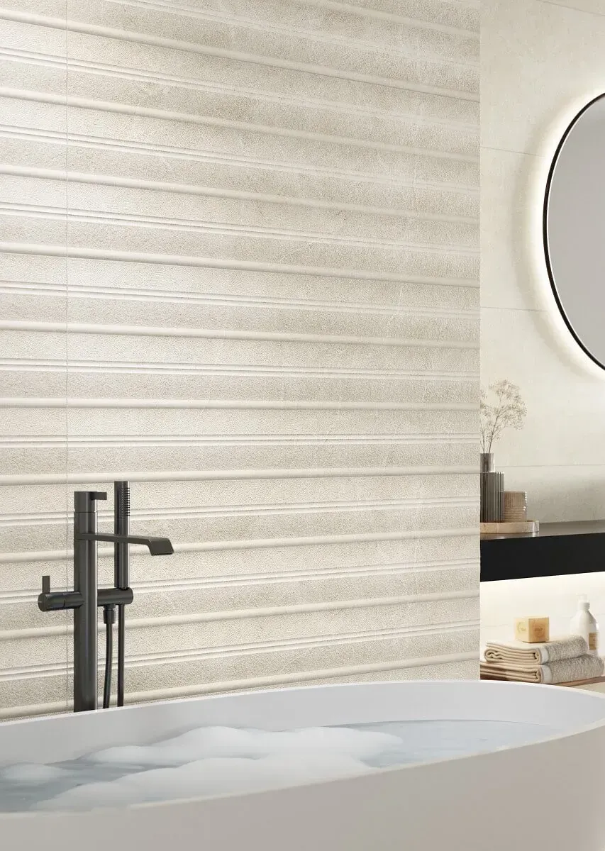 Baño con pared de azulejos beige texturizados, grifo negro y bañera blanca. Espejo redondo y elementos decorativos visibles.