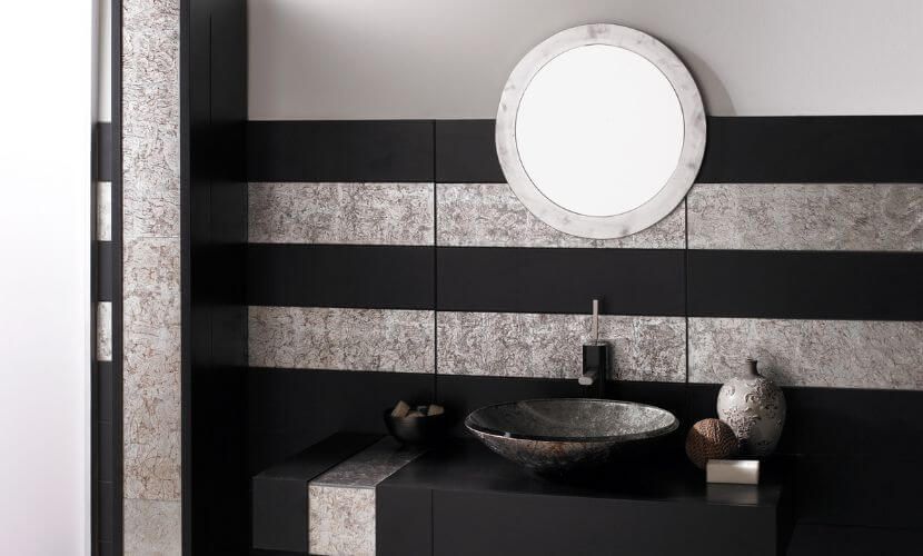 Baño con azulejos de pared con rayas negras y plateadas, espejo redondo, tocador oscuro y lavabo tipo vasija.