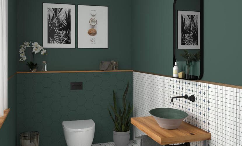 Baño con paredes verdes, cenefa de azulejos blancos, tocador de madera, grifería negra e inodoro. Decoración artística y vegetal.