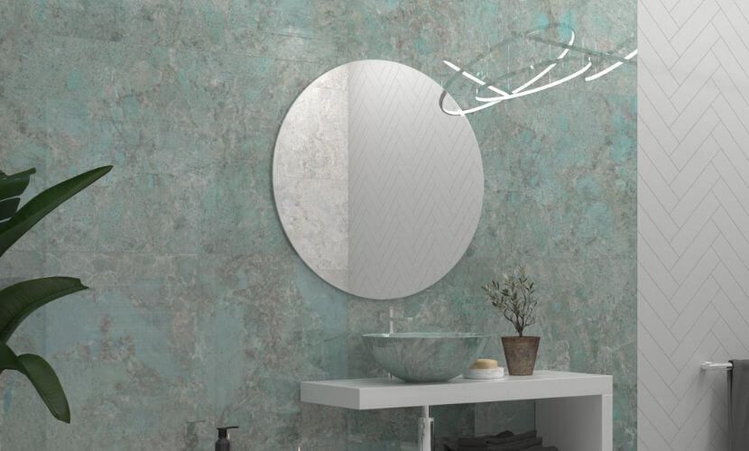 Baño con espejo redondo, pared con textura verde azulado, tocador blanco y lámpara moderna.