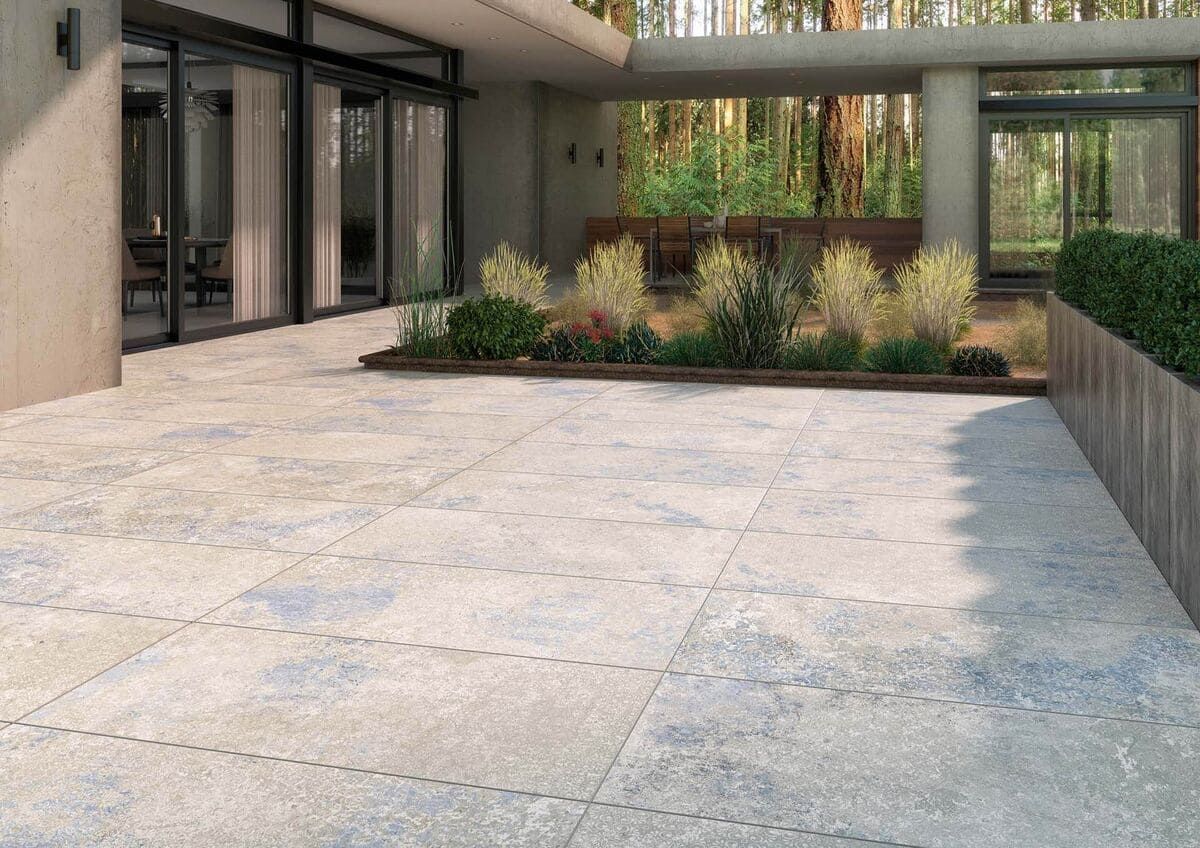 Patio exterior con suelo de piedra azul claro y gris, cama de jardín y casa moderna con puertas de cristal.