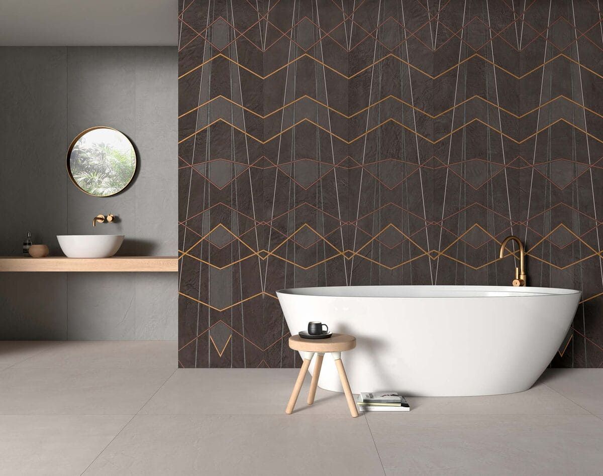 Baño con pared decorativa con patrón oscuro, bañera blanca, accesorios dorados y espejo redondo.