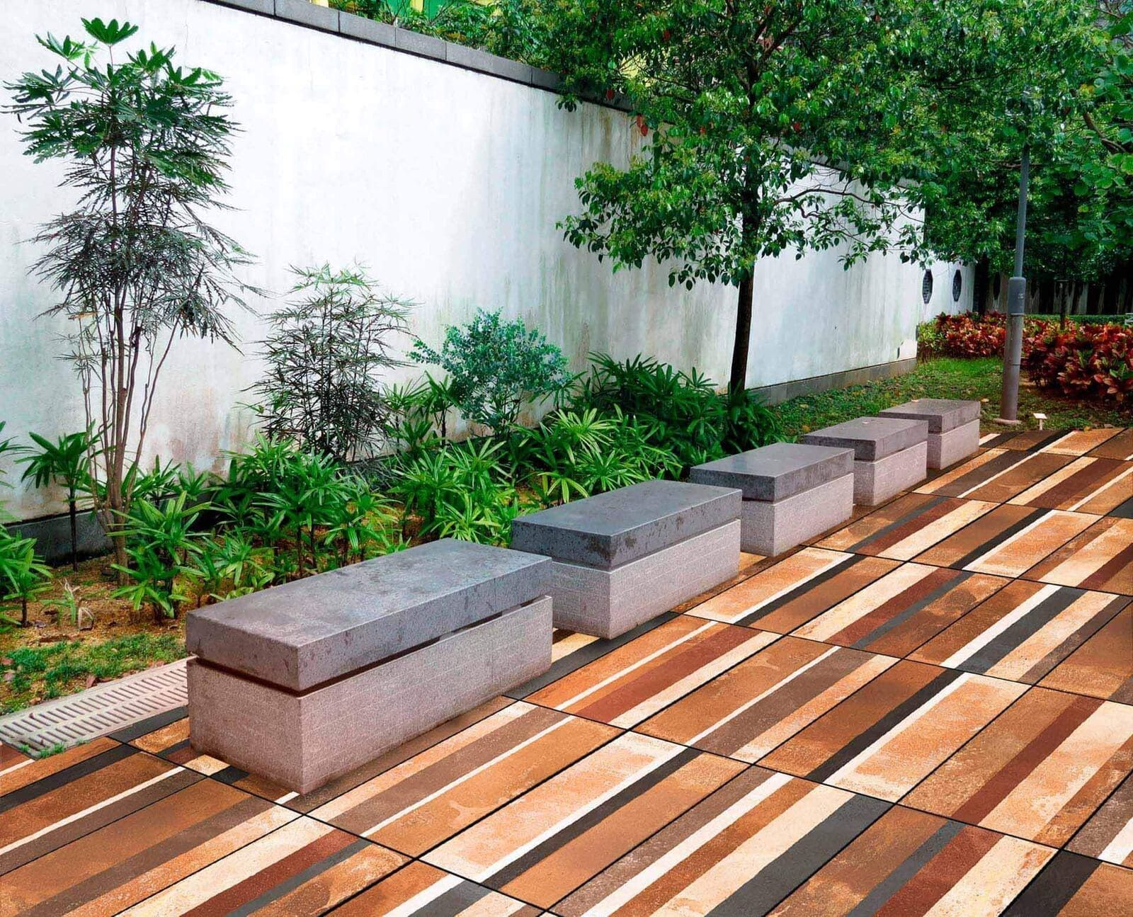 Fila de bancos rectangulares de piedra sobre una pasarela de baldosas con aspecto de madera al lado de una pared con jardinería.