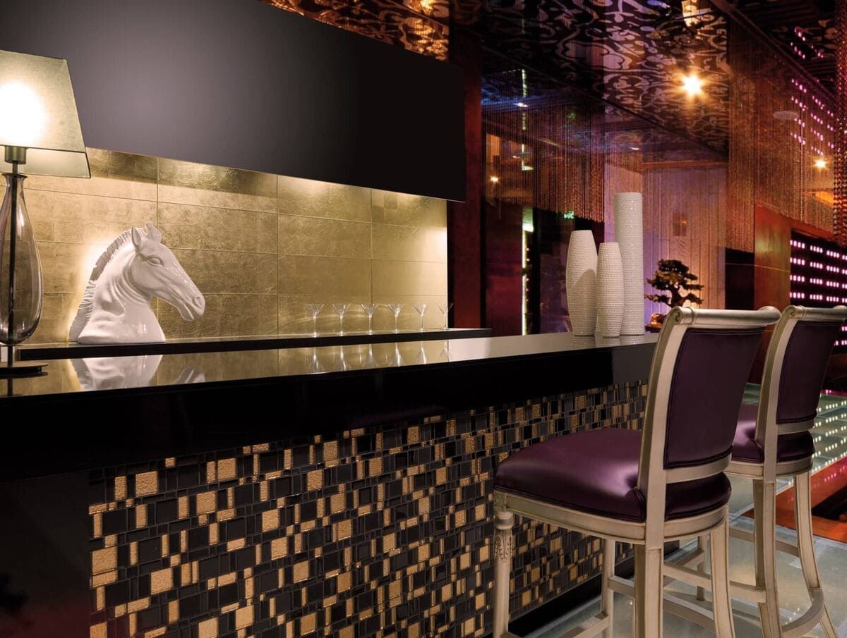 Interior de bar con taburetes, frente de mosaico, pared con detalles dorados y una escultura de caballo.