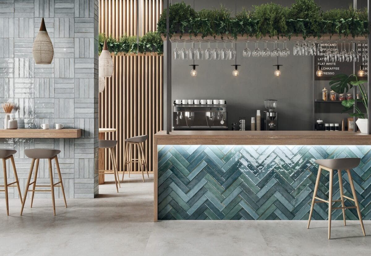 Interior de bar con detalles de madera, frente de barra con azulejos azules, taburetes y plantas en lo alto.