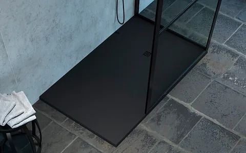 Plato de ducha rectangular negro, mampara de cristal, suelo de baldosas, toallas.