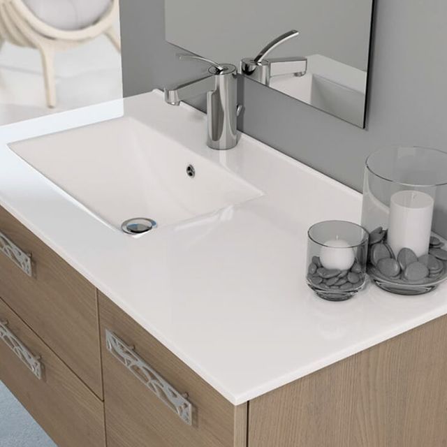 Lavabo blanco sobre un mueble de madera, con grifo y espejo. Junto al lavabo hay velas y piedras decorativas.