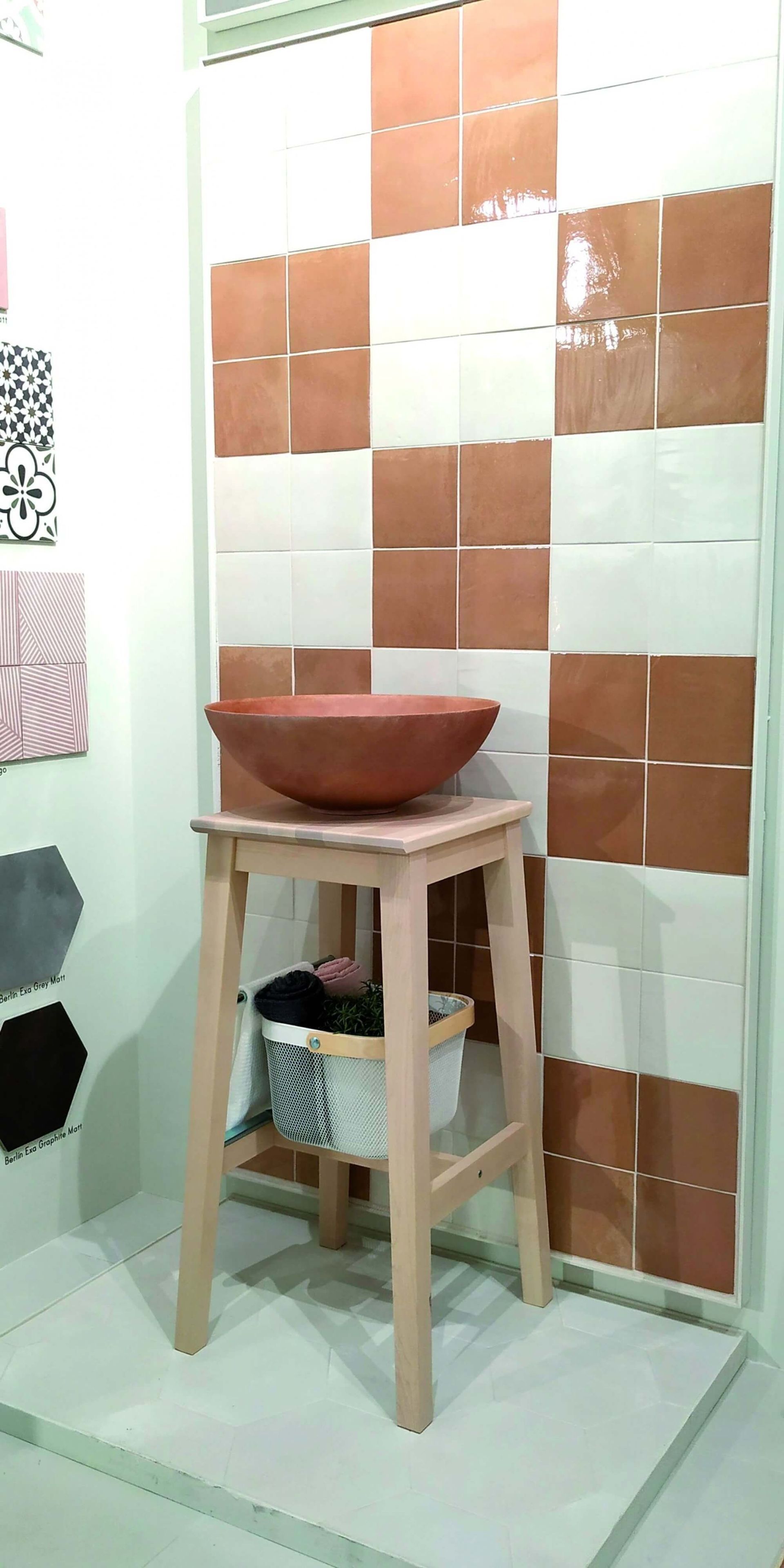 Un expositor de baño con una pared de azulejos a cuadros, un taburete de madera que sostiene un lavabo y una cesta de tela.