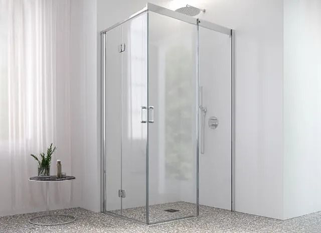 Ducha esquinera con puertas de cristal transparente y marco plateado. Paredes blancas y suelo de guijarros.