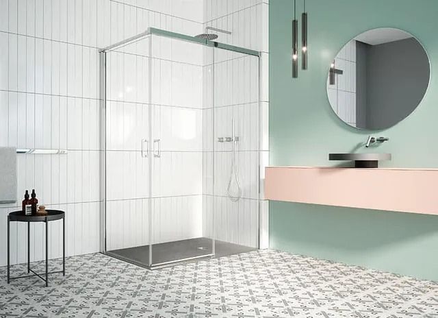 Baño con ducha de cristal, lavabo rosa y suelo estampado. Azulejos verticales blancos en la pared.